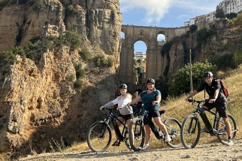 Three cyclists on a dirt path with a rocky cliff and an ancient aqueduct in the background. 2