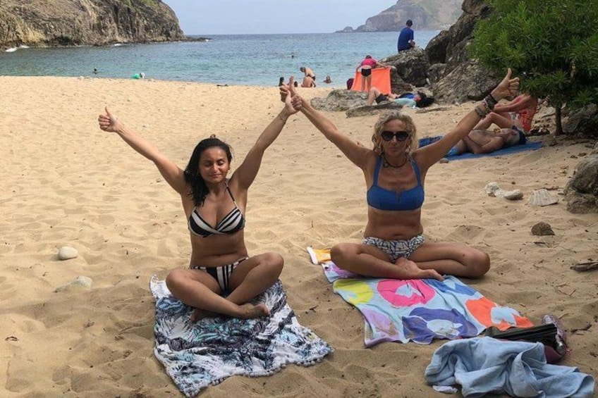 Two people sitting on the beach with their arms raised, surrounded by beach towels and a rocky coastline. 5