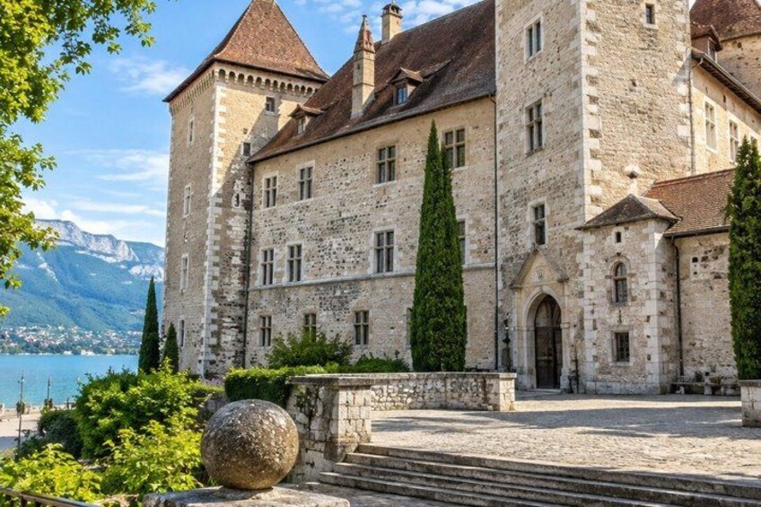 A historic stone building with a tower, surrounded by greenery and a body of water in the background. 5