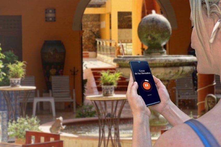 A person holding a smartphone with a music app open, outdoors with a fountain, chairs, and potted plants. 5