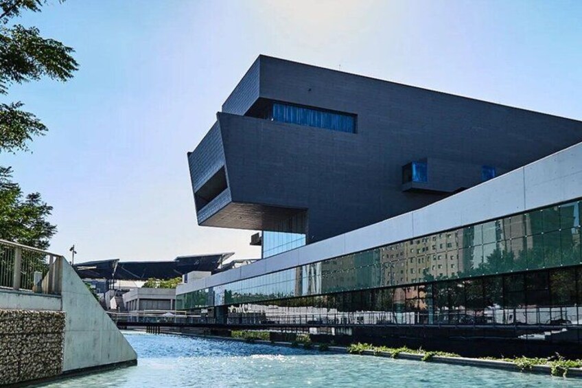 A modern building with a unique design, a river, and a bridge in the background. 2