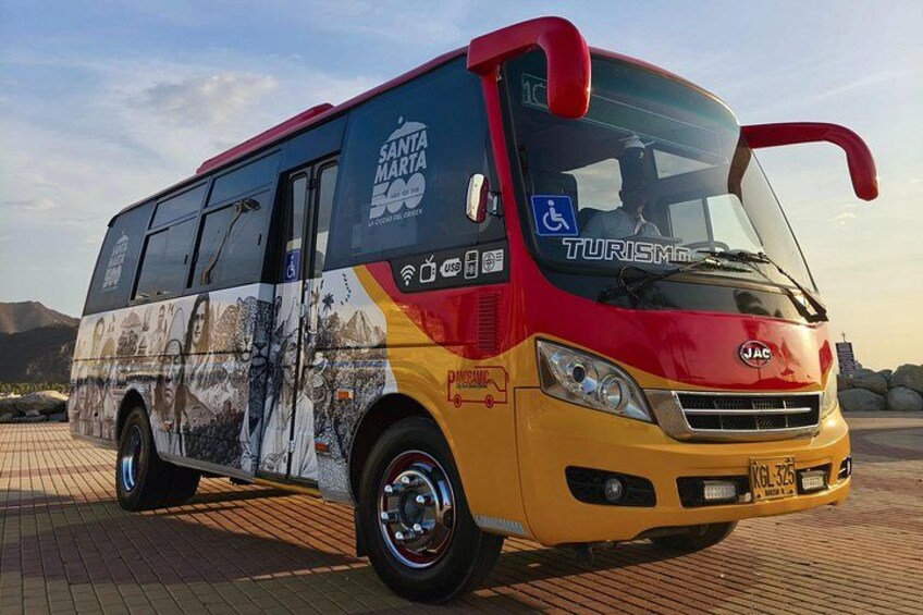 A red and yellow bus with 'Santa Marta' and 'Turismo' on the side, parked on a paved area with mountains in the background. 1