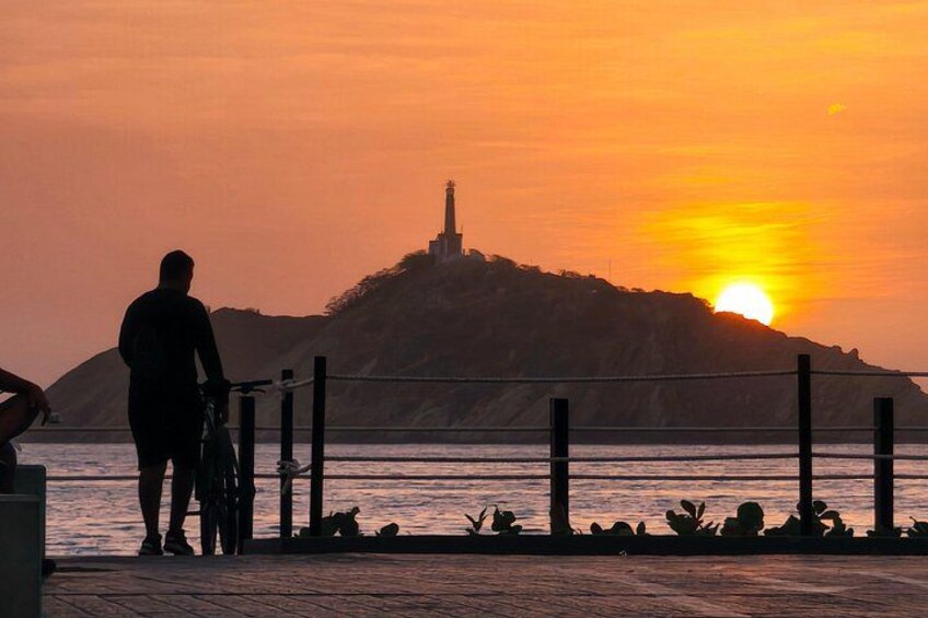 A sunset over a body of water with a silhouette of a person and a bicycle, a fence, and a distant hill with a structure. 5