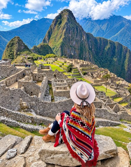 Eine Person sitzt auf einem Felsen und blickt auf die Ruinen von Machu Picchu mit Bergen im Hintergrund. 1