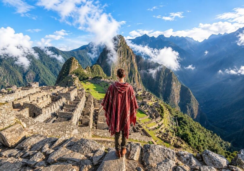 Eine Person steht auf Steinstufen mit Blick auf Machu Picchu, im Hintergrund Berge und Wolken. 3