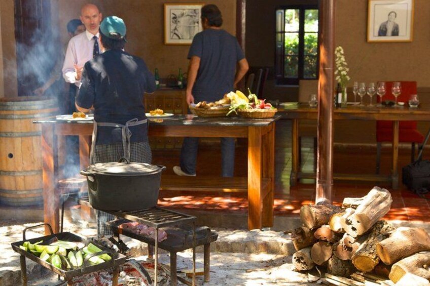A rustic outdoor dining area with a large pot on a stove, a table with food, and a wooden barrel. 3