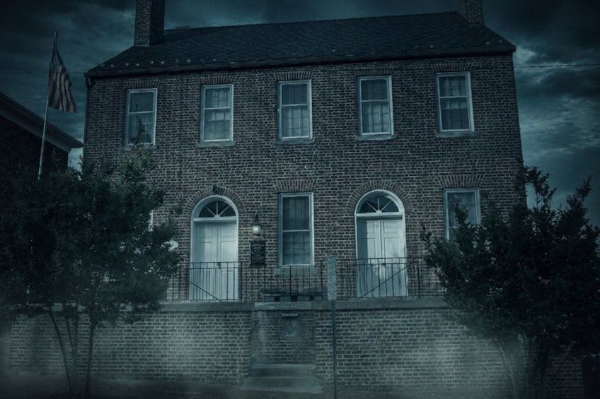 A brick building with arched windows and a white door, surrounded by trees and a fence. 2