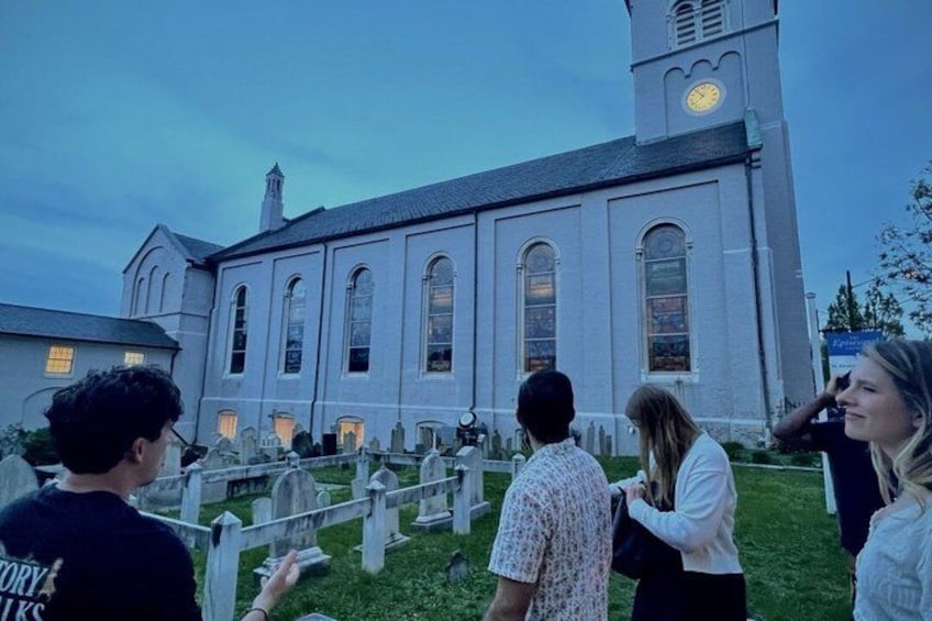 A church with a clock tower, a cemetery with headstones, and people standing in front. 3