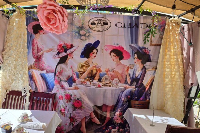 A vintage-themed outdoor dining area with a mural of women in period attire, a table set for tea, and a tent overhead. 2