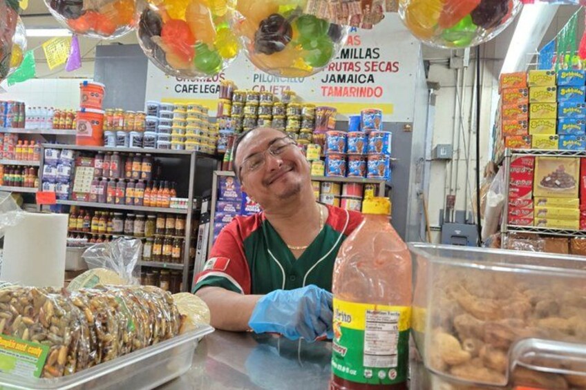 A person in a red and green uniform behind a counter with various food items and a sign in the background. 1
