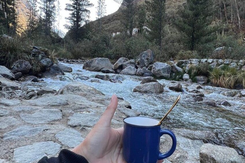 A hand holding a blue mug with a spoon, overlooking a rocky riverbed with trees in the background. 2