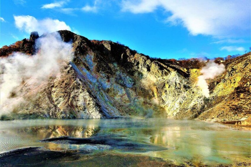 A geothermal area with steam rising from the ground, a reflective water surface, and a mountainous landscape. 1