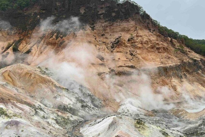 A geothermal area with steam rising from the ground, surrounded by rocky terrain and sparse vegetation. 3
