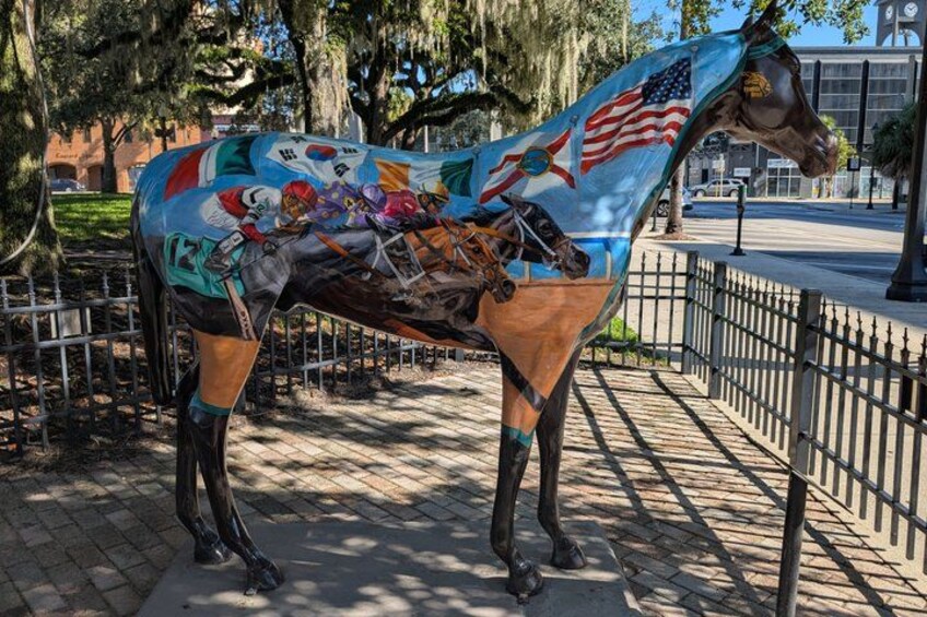 A horse statue with a colorful mural, surrounded by a fence and trees. 5