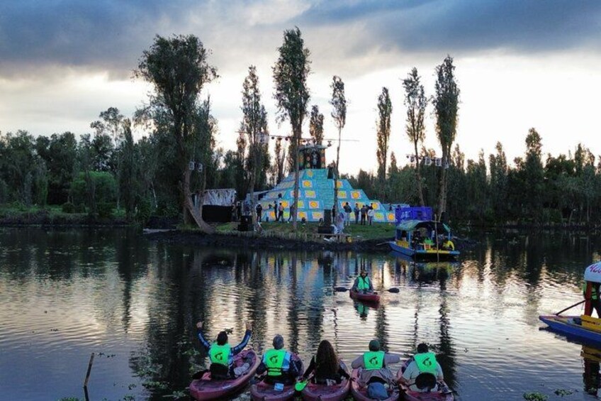 A group of people in canoes on a calm lake, a colorful structure in the background, and trees lining the shore. 2