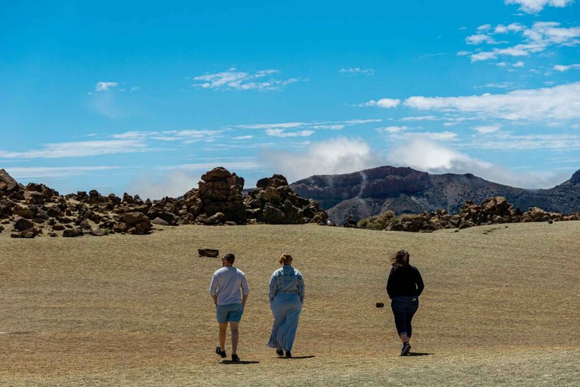 Trois personnes marchent sur un terrain aride et rocheux, avec un paysage montagneux en arrière-plan et un ciel dégagé. 3