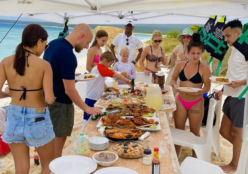 A group of people dining outdoors with a table full of food and drinks, beach chairs, and a tent in the background. 5
