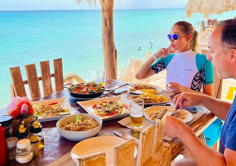 A wooden table with various dishes and drinks, overlooking a turquoise sea. 1