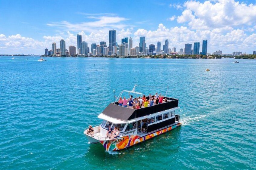 A double-deck boat with passengers on a clear day, cruising on turquoise water with a city skyline in the background. 1
