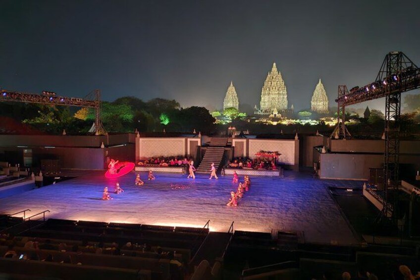 A nighttime outdoor performance with dancers on a stage, a large illuminated structure in the background, and a river. 1