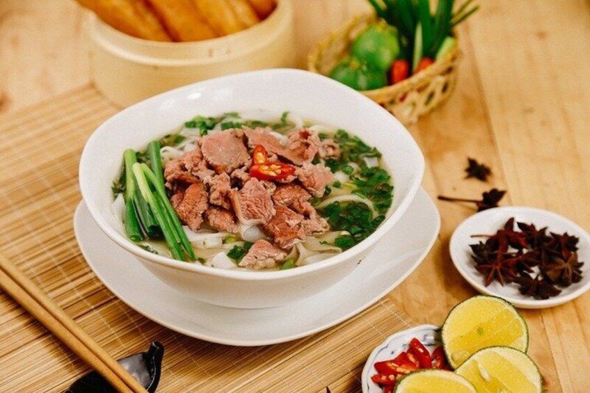A bowl of soup with meat and vegetables, a plate of sliced lemons, and a small bowl of chili peppers on a wooden table. 3