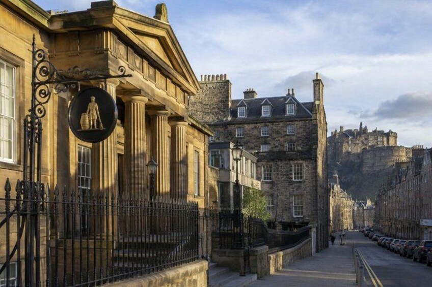 A historic street with stone buildings, a prominent castle in the background, and a black metal fence. 2