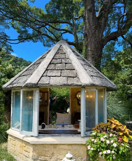 A stone gazebo with a pointed roof, surrounded by lush greenery and a large tree. 4