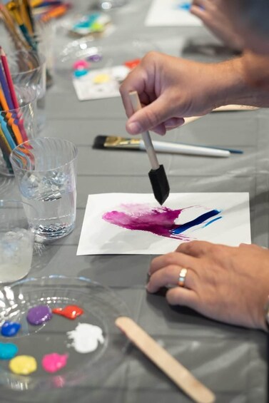 Une personne est en train de peindre sur une feuille de papier blanche à l’aide d’un pinceau, entourée de pots de peinture et d’autres pinceaux sur une table. 3