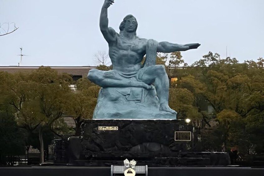 A large bronze statue of a seated man with one arm raised, set on a pedestal in a park with trees and buildings in the background. 2