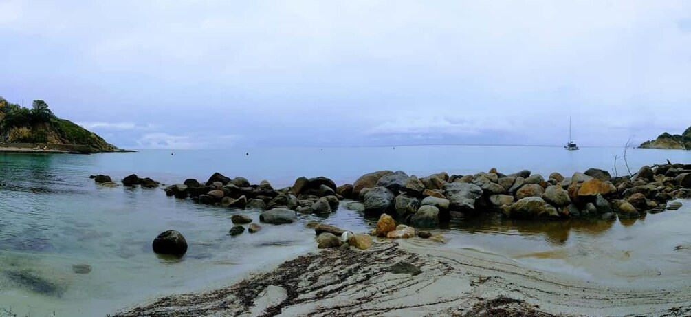 A rocky shoreline with clear water, a sailboat in the distance, and a hilly landscape in the background. 3