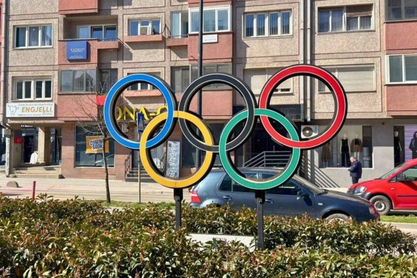 Olympic rings, a blue car, and a red car parked in front of a building with balconies. 4