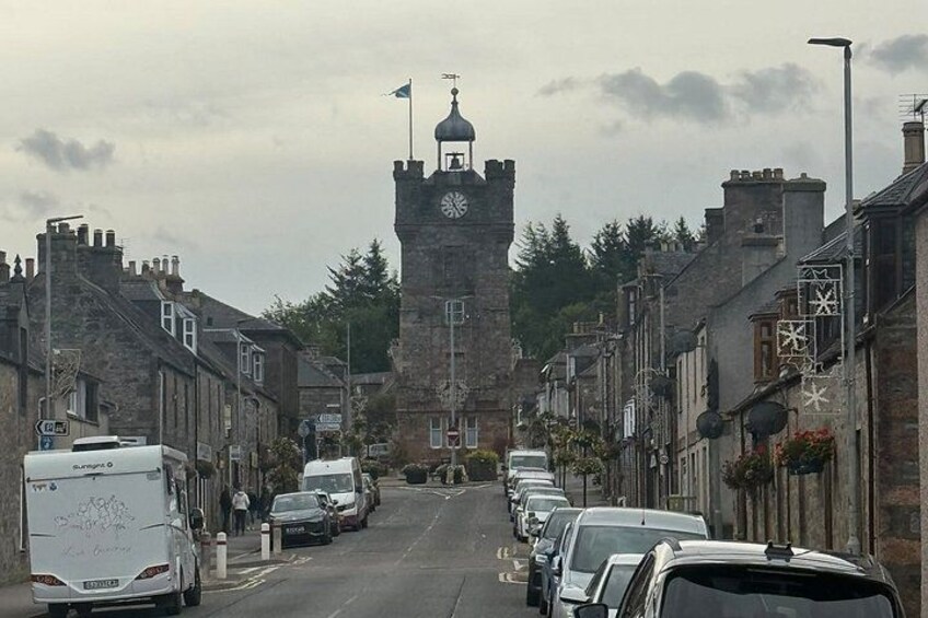 A street with parked cars, a clock tower, and stone buildings. 2