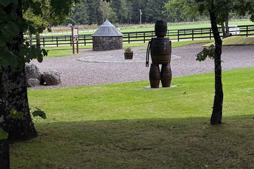 A sculpture of a human figure made from barrels, a stone structure, and a fenced area with a gravel path. 5
