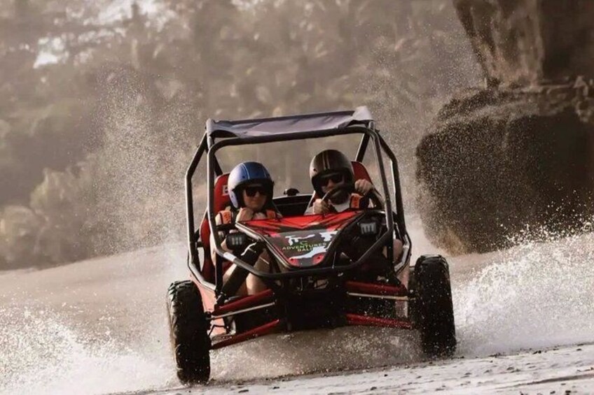 Two people in a red ATV driving on a dirt road with dust being kicked up. 1