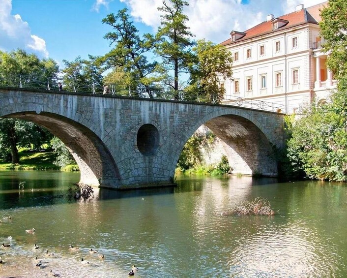 Un pont de pierre enjambe une rivière paisible où nagent des canards, le tout étant entouré d’arbres et d’un bâtiment dont le toit est rouge. 1
