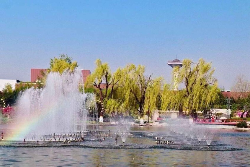A fountain with water jets, a rainbow, and willow trees in a park. 2