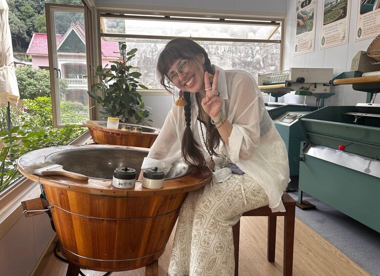 Eine Person sitzt in einer Holzbadewanne mit Blick durch ein Fenster auf ein Haus und Grünflächen. 4