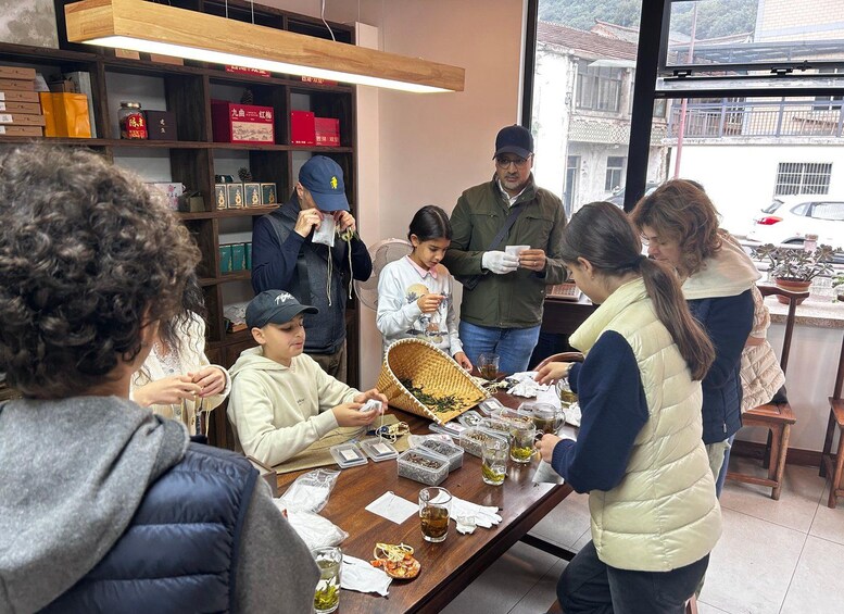 Eine Gruppe von Menschen nimmt an einer Teezeremonie in einem traditionellen Ambiente teil, mit Teekästen an Regalen und einem Holztisch mit Teeutensilien. 3