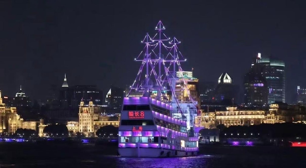 Een fel verlichte dubbeldekkerboot met een piramidevormige constructie bovenop, die op een rivier vaart met een stadsachtergrond. 3