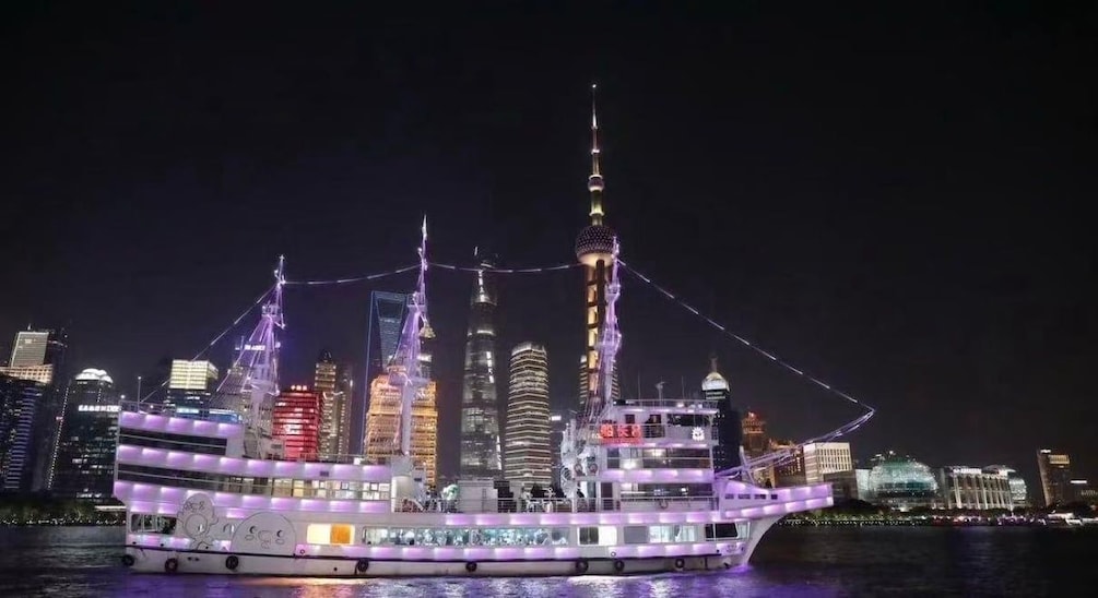 Een helder verlicht cruiseschip met een modern ontwerp, varend over water met een stadsachtergrond. 1