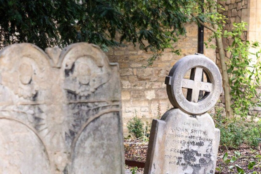 A gravestone with a cross symbol and inscriptions, set against a stone wall and greenery. 2