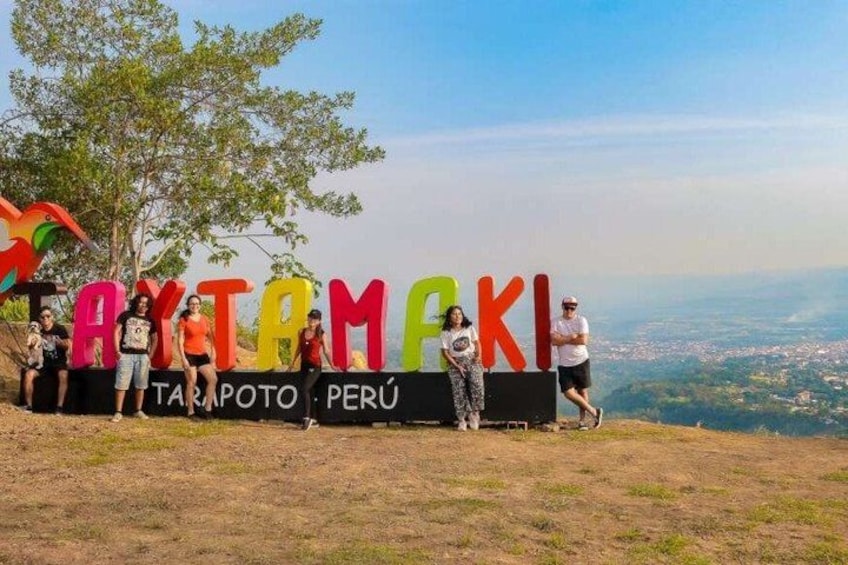 A sign with colorful letters spelling 'TAYTAMAKI' and 'TARAPOTO PERU' on a hillside with a view of the city below. 1