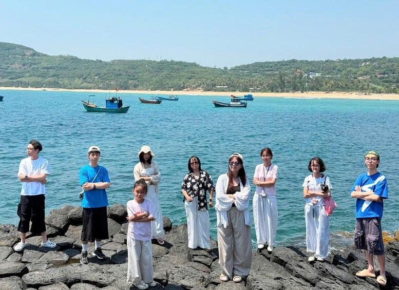 A group of people standing on rocks by a calm sea with boats in the background. 3