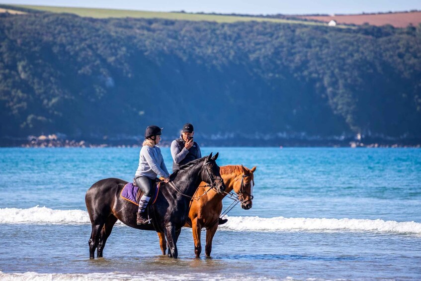 Two people riding horses along a beach with waves and a forested hill in the background. 1