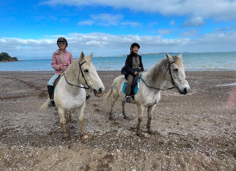 Two people riding horses on a pebbly beach with a body of water and distant land in the background. 2