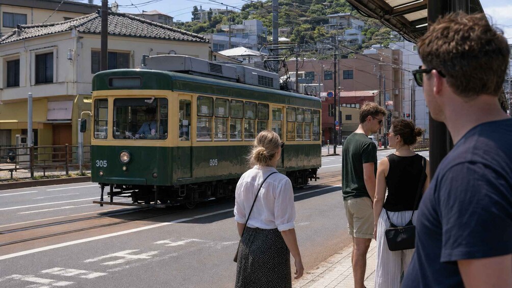 A green and yellow tram on tracks, passengers waiting on the platform, and buildings in the background. 3