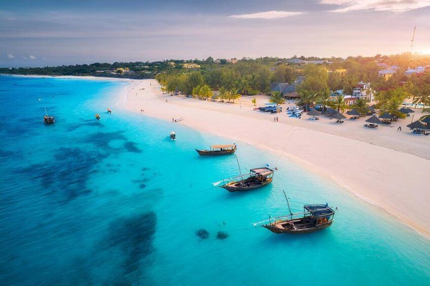 A beach with clear turquoise water, several boats anchored near the shore, and a line of houses in the background. 3