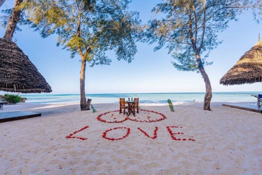 A beach with a heart-shaped LOVE symbol drawn in the sand, palm trees, and thatched umbrellas. 3