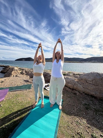 Deux personnes pratiquent le yoga sur un tapis bleu, au bord d’une côte rocheuse, avec un plan d’eau et des collines en arrière-plan. 3