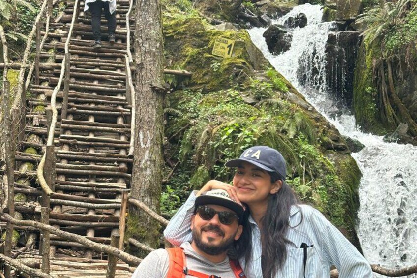 A wooden bridge over a waterfall with moss-covered rocks and a person standing on it. 1
